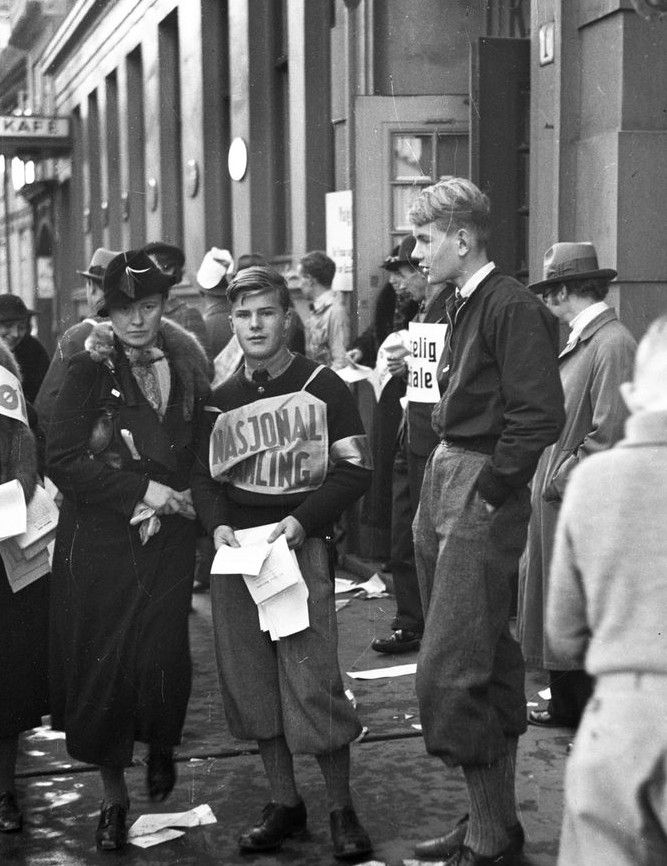 To unge menn i nikkers og jakker står sammen med en dame med hatt og pels. En av de unge mennene har festet et skilt til brystet der det står Nasjonal Samling. Han holder noen løpesedler i hånden. Svart-hvitt foto.