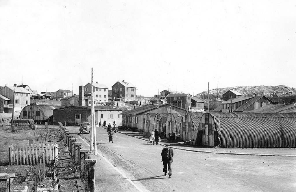 Flere toetasjes hus med saltak ligger på høydedraget i bakre det av bildet. I fremre del av bildet går flere mennesker langs ei gate hvor det også er en bil. I gata er det flere lange, lave skur med rundbuede bølgeblekktak som går helt ned til bakken. Foto. 