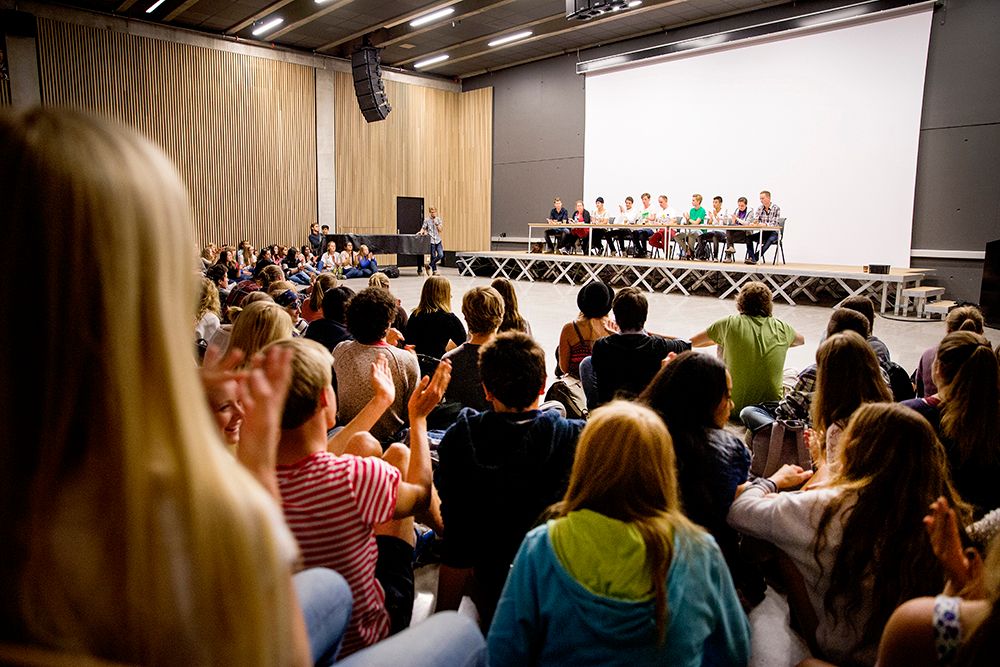Skoledebatt. Det sitter ti personer på rekke bak et bord foran et hvitt lerret. En person står litt på siden og leder debatten. I forgrunnen sitter mange ungdommer i publikum. Foto. 