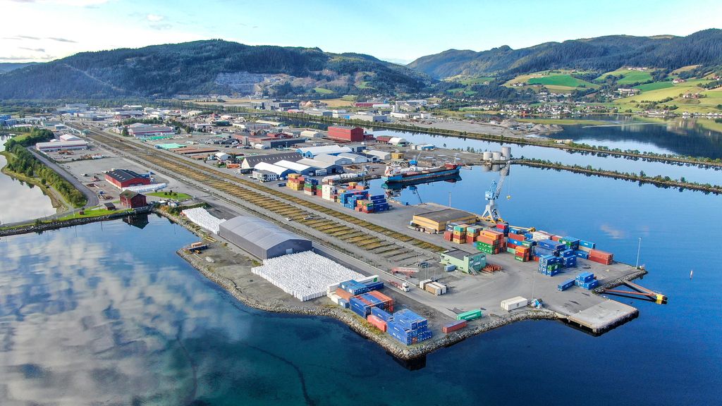 Havneområde med bebyggelse, åkre og åser i bakgrunnen. Ytterst på havna er ulike konteinere stablet opp. Foto.