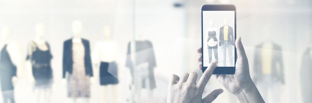 Hender som holder en mobiltelefon. På mobiltelefonen ser vi at det blir tatt bilde av en vindusutstilling. Foto.