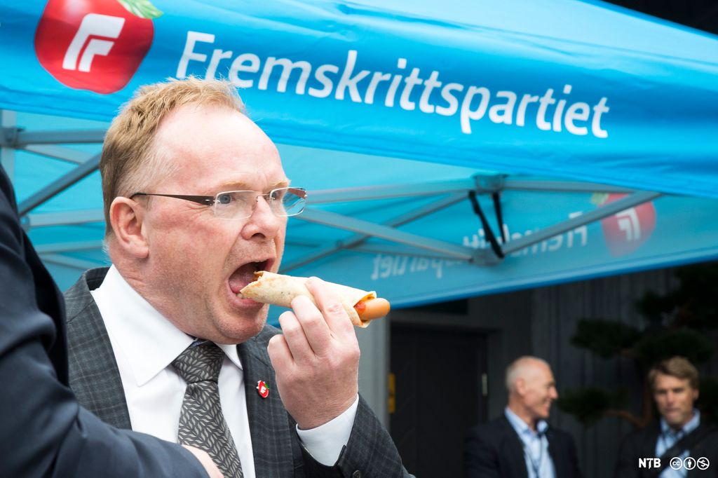 Mann spiser pølse i lompe under en parasoll med Fremskrittspartiets logo. Foto.