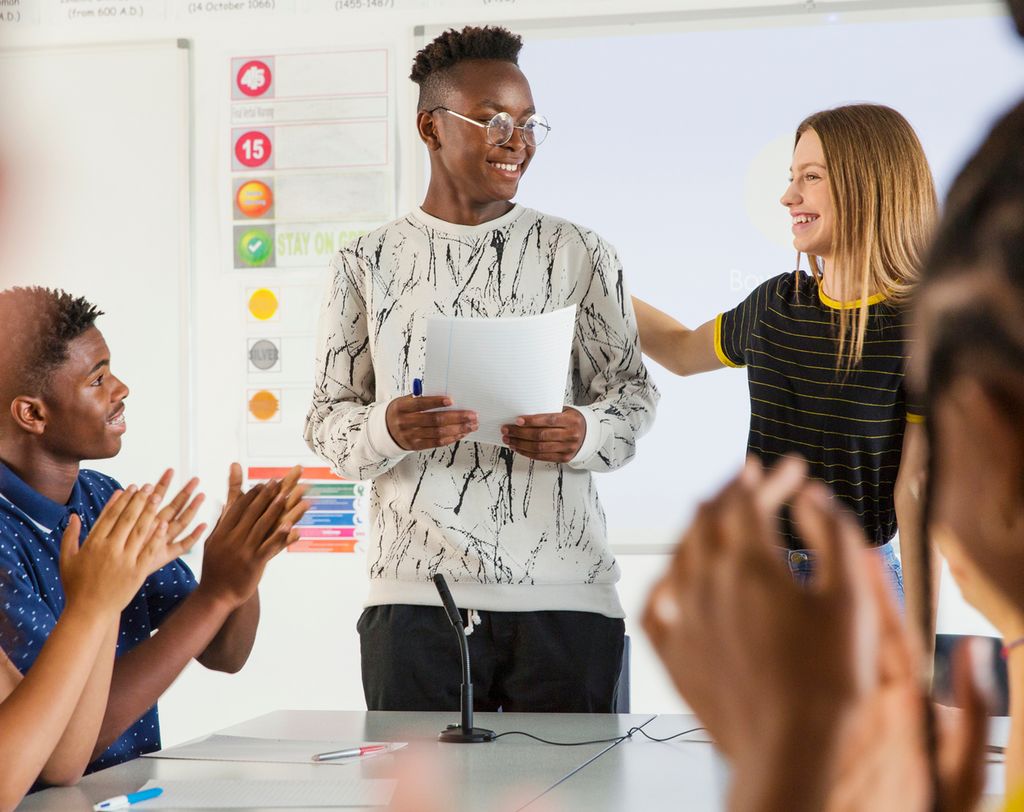 One pupil standing at the board with a teacher. The other pupils are gathered around a table. The other pupils clap. Photo.