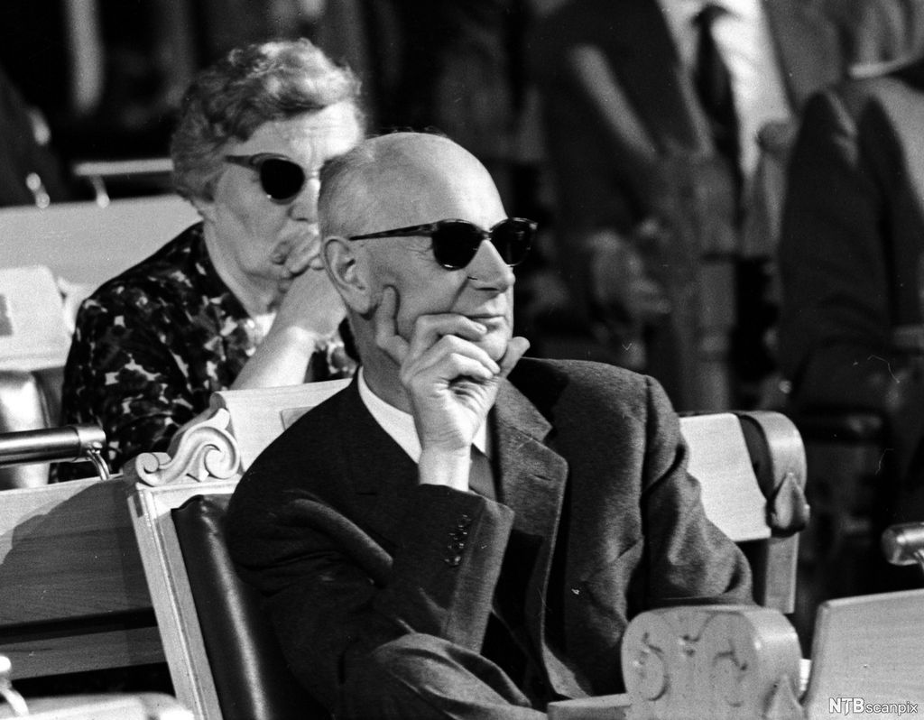 Einar Gerhardsen sitter i Stortingets sal med solbriller på og lytter. Foto.