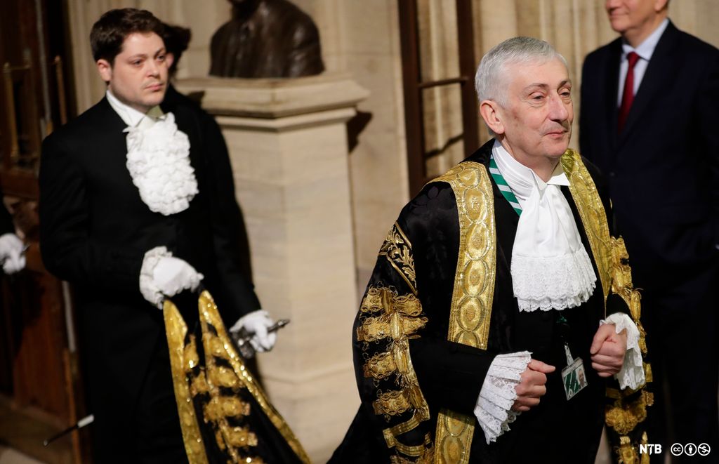 Photo: We see a man with grey hair dressed in elaborate old-fashioned clothes. He is wearing a long, black cloak, edged with broad gold ribbons. His shirt has a lot of lace. On his stomach, we can see a plastic ID card hanging from a green and white lanyard. A man is walking behind him, holding up his cloak. The man is also dressed in old-fashioned clothes; he wears a lace collar and lace cuffs and white gloves. 
