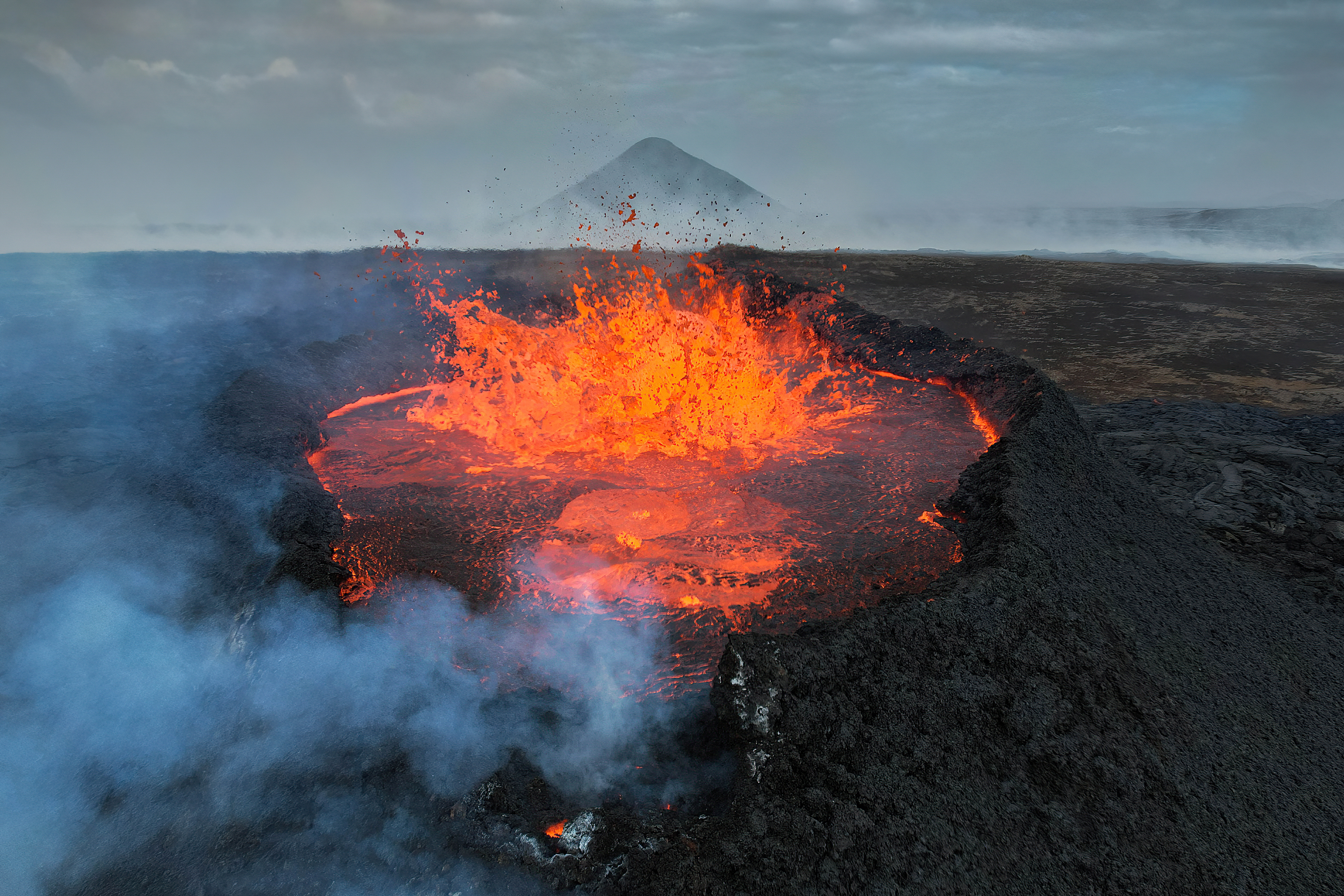 Glødende lava som spruter opp fra en vulkan som omgitt av mørk stein og røyk. Foto.