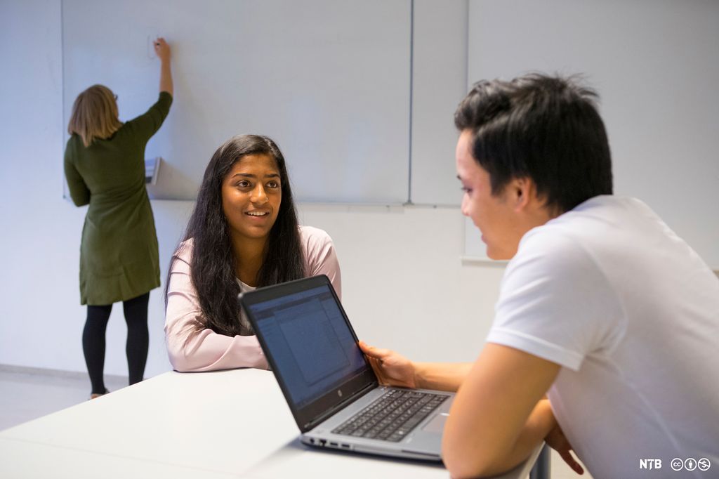 Jente og gutt sitter med en PC i forgrunnen. Lærer skriver på tavla i bakgrunnen. Foto. 