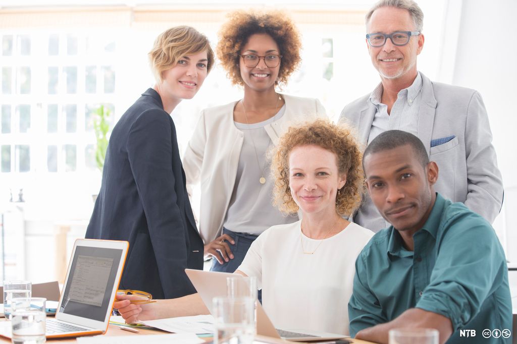 Gruppe med fem personer i et kontorlandskap. To av dem sitter ved et arbeidsbord, de tre andre står bak dem. Foto.