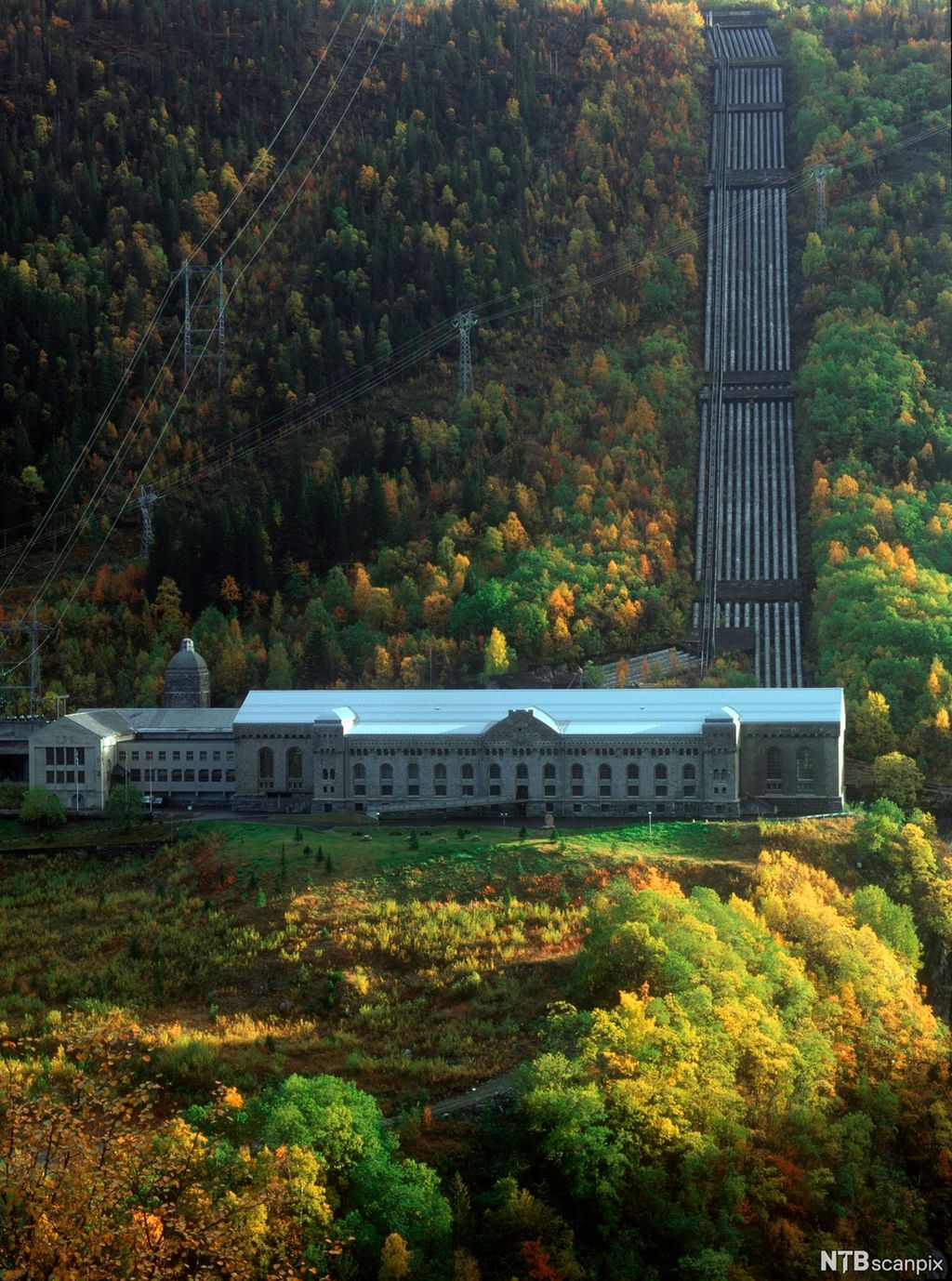 Vannkraftstasjon i bratt fjellside. Rørgate fører vann inn i anlegget, og master bærer høyspentledninger som transporterer elektrisiteten ut. Foto.