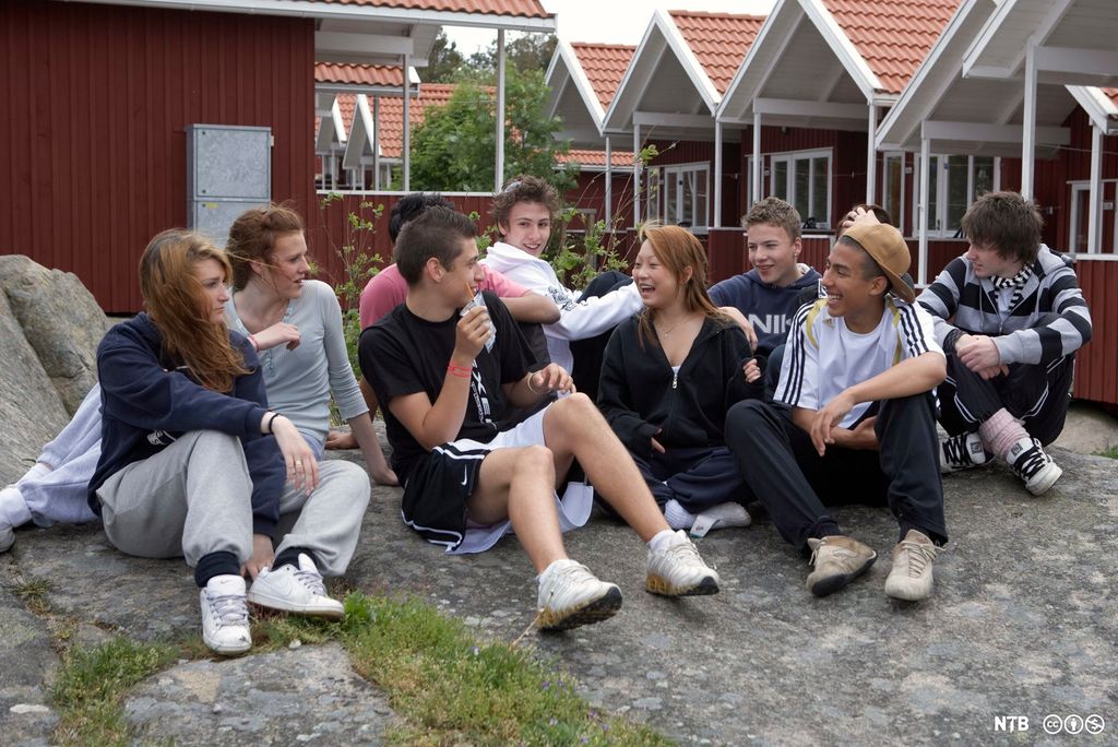 En gjeng ungdommer sitter sammen på bakken ute. De snakker sammen og ler. Noen har shorts og T-skjorte på. I bakgrunnen er det rekker med små hus eller hytter. Foto.
