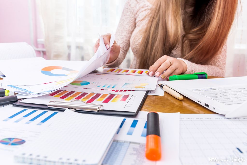 Kvinne jobber med bærbar datamaskin og flere ark med grafiske framstillinger. Foto.