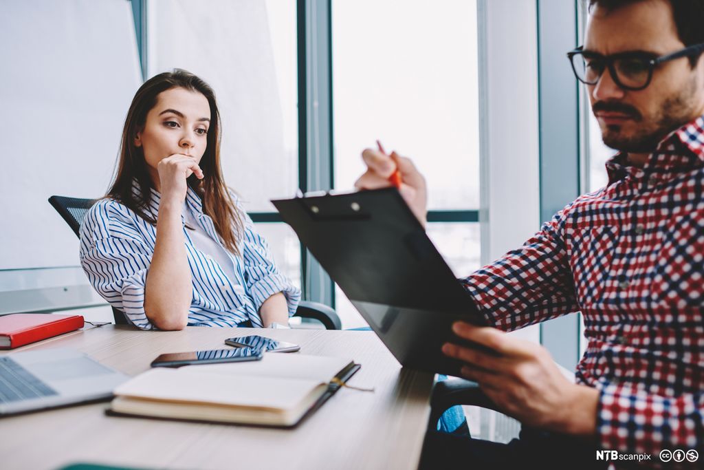 En mann og en kvinne ved et bord med bøker, laptop og mobiler. Mannen ser alvorlig på noe på et ordrebrett. Foto.