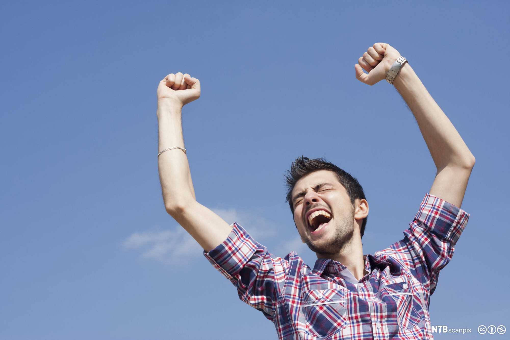 Glad ung mann som løfter hendene i været mot en blå himmel. Han kniper sammen øynene og åpner munnen som om han jubler. Han har rutete skjorte med oppbrettede ermer. Foto.