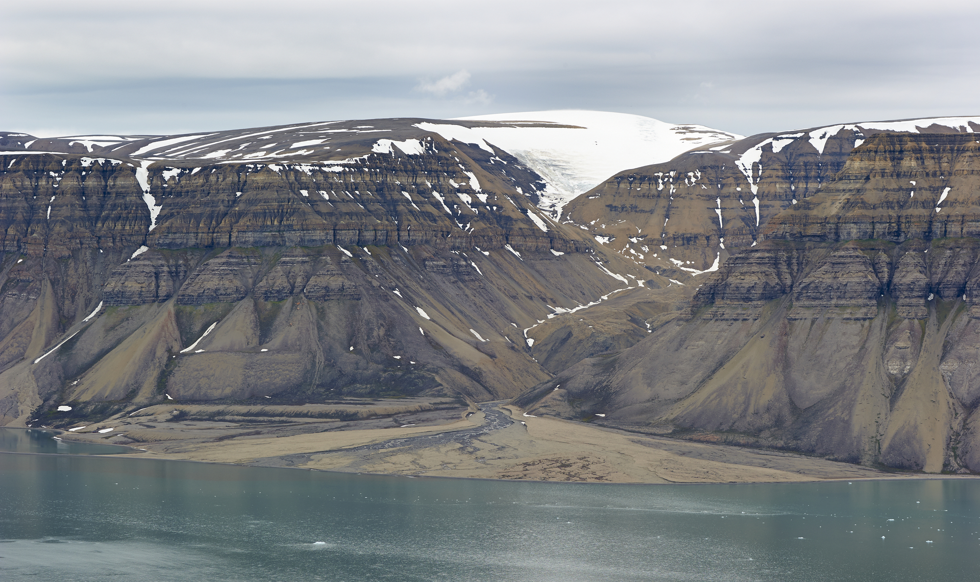Elv renn i eit søkk i eit fjell med horisontale striper og nedover mot ein fjord. Det ligg snø på toppen av fjellet. Der elva renn ut i fjorden, er det avsett lausmassar på ein slik måte at dei dannar ein brun trekant. Foto.