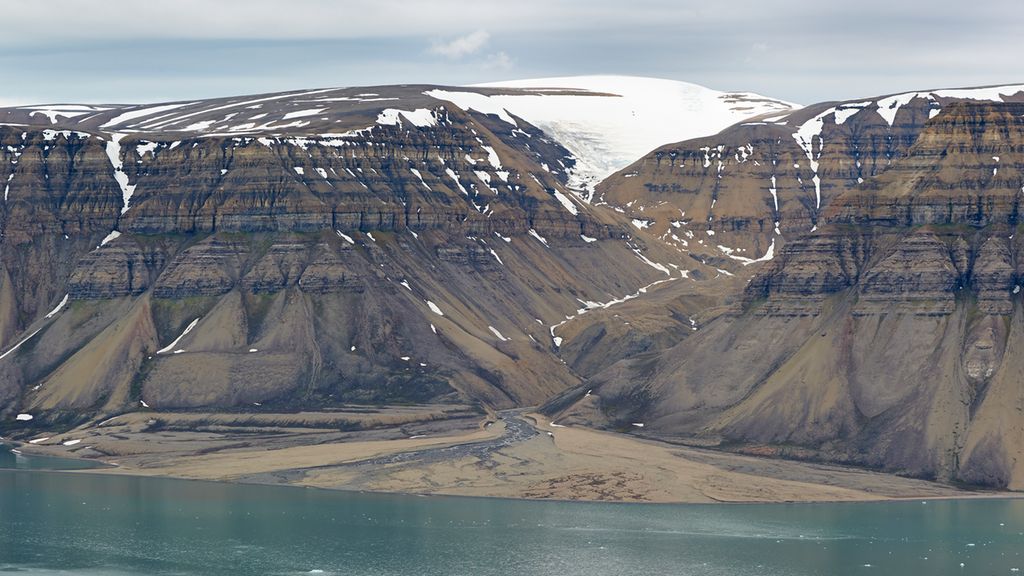 Elv renn i eit søkk i eit fjell med horisontale striper og nedover mot ein fjord. Det ligg snø på toppen av fjellet. Der elva renn ut i fjorden, er det avsett lausmassar på ein slik måte at dei dannar ein brun trekant. Foto.