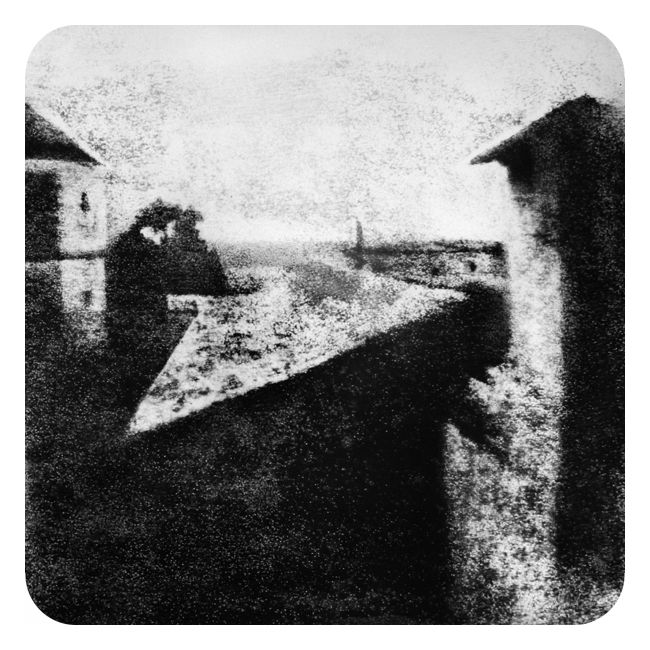 Svært uskarpt svart-hvitt-fotografi som viser noen hustak. Landskapet bakenfor er flatt. Det er vanskelig å si om skyggene i bakgrunnen er trær eller bygninger. Himmelen er høy og lys, bildet er tydeligvis tatt på dagtid. 