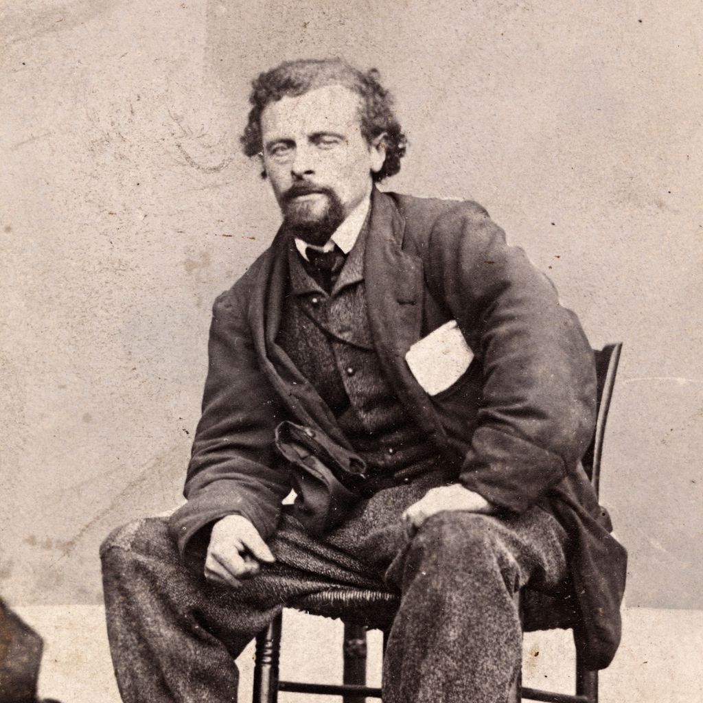 Portrett av Marcus Thrane ca. 1863. Han sitter bredbeint og litt framoverlent på en stol. Han er uvørent kledd, buksa er oppbretta og har lapp på kneet. Han har noen papirer i brystlomma. Foto.