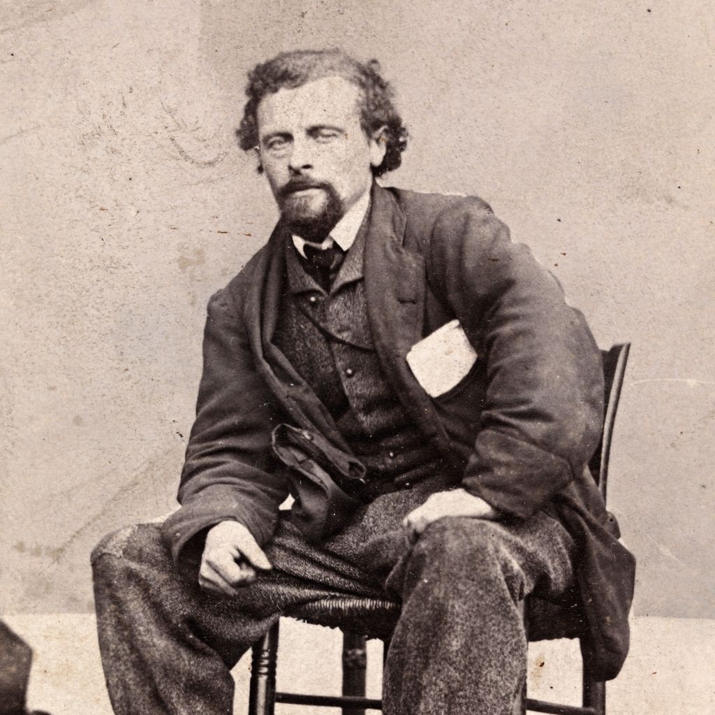 Portrett av Marcus Thrane ca. 1863. Han sit breibeint og litt framoverlent på ein stol. Han er uvørdent kledd, buksa er oppbretta og har lapp på kneet. Han har nokre papir i brystlomma. Foto.