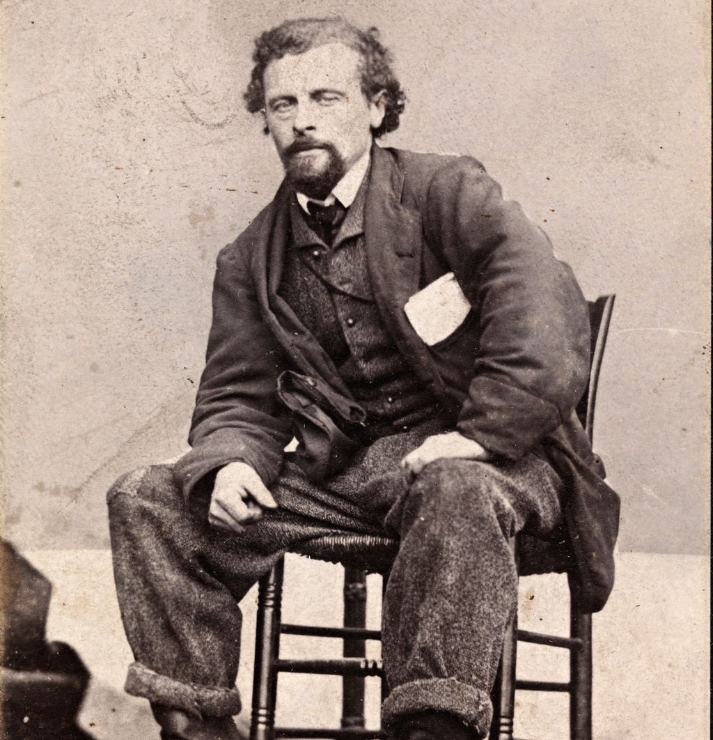 Portrett av Marcus Thrane ca. 1863. Han sitter bredbeint og litt framoverlent på en stol. Han er uvørent kledd, buksa er oppbretta og har lapp på kneet. Han har noen papirer i brystlomma. Foto.