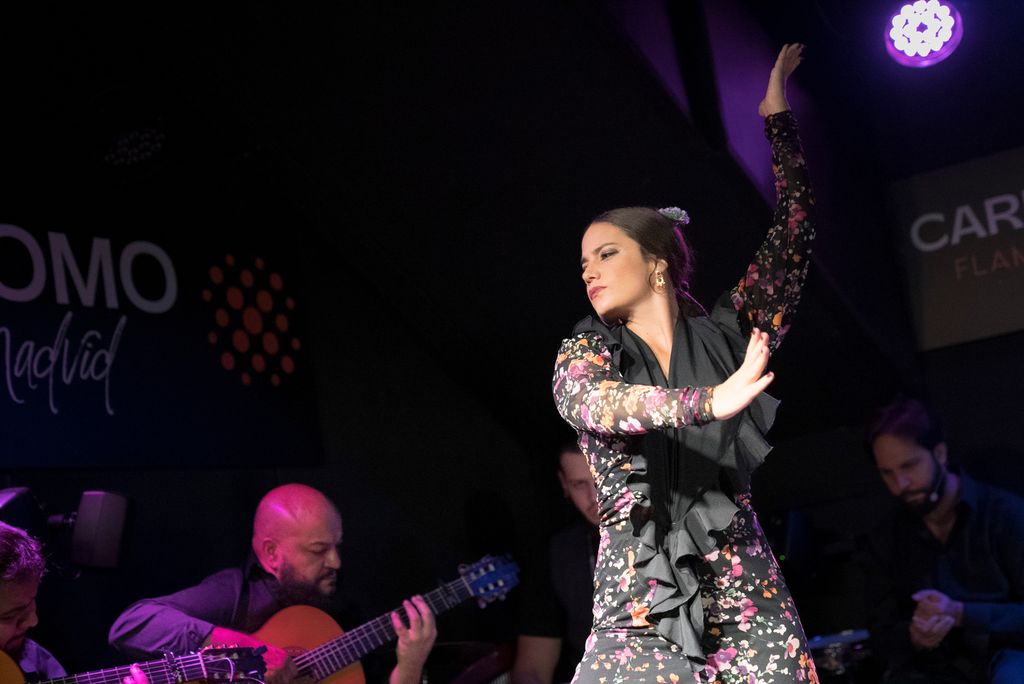 En kvinne i scenelys danser flamenco. Det er musikere i bakgrunnen. Foto.