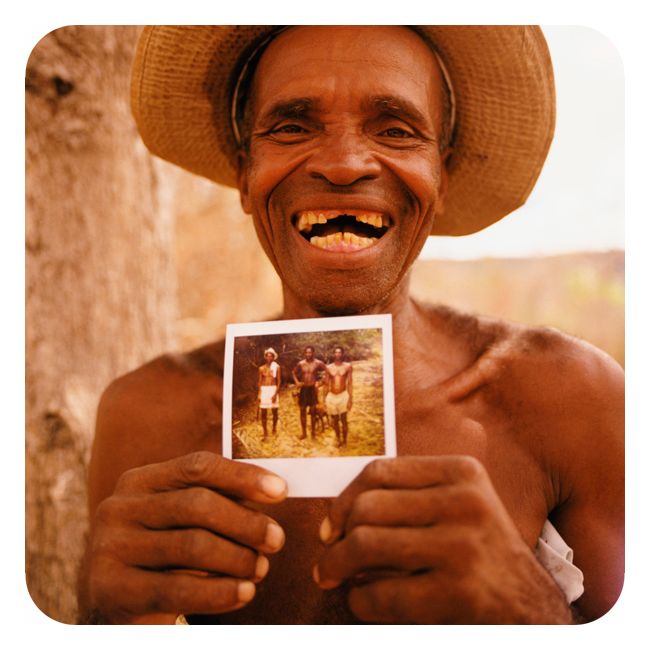 En mann smiler hjertelig mot et kamera. Han har bar overkropp og en hatt på hodet, og han har mistet fortennene. I hendene holder han et fotografi som viser tre menn. Det kan se ut som om han selv er en av disse mennene. 