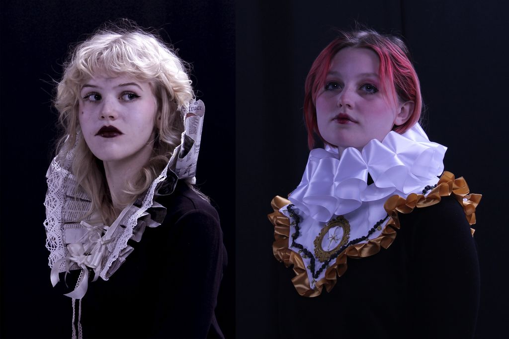 To unge damer har på seg en skulpturell krage laget i tekstil og i en kombinasjon av tekstil og avispapir. Portrettene er lyssatt mot en mørk bakgrunn. Dette skaper en kontrast som gir bildet en dramatisk stemning. Foto.