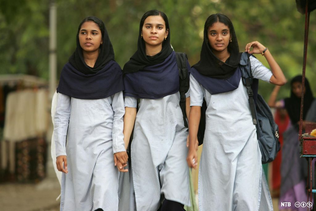Three Indian girls walking hand in hand, They are dressed in similar dresses and scarves, probably a school uniform. They look serious and focused. Photo.