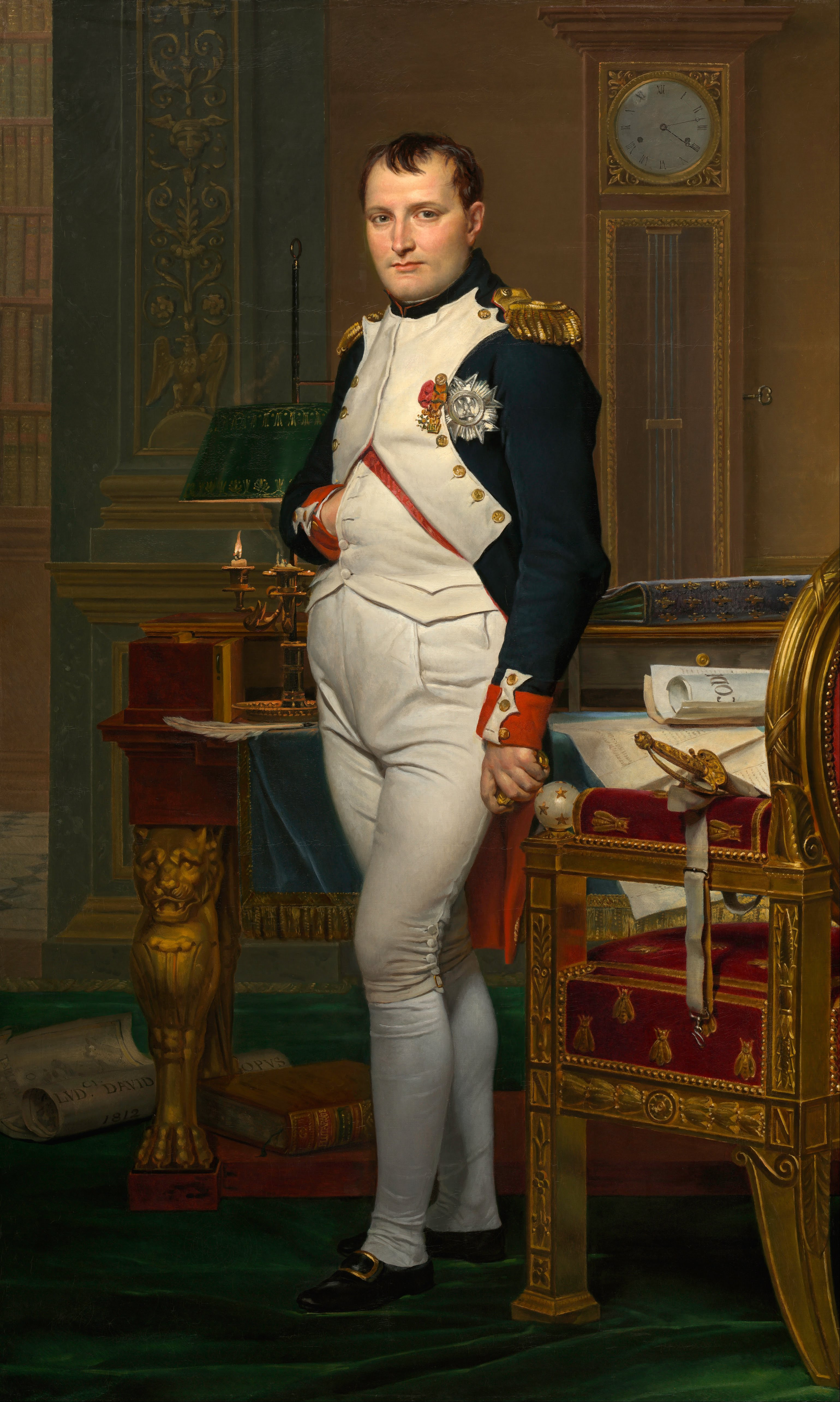 Napoleon i uniform i sitt kontor. Han ser mot betrakteren og har hånden karakteristisk stukket inn i jakken foran. Jakken er utstyrt med medaljer og gull på skuldrene. Maleri. 