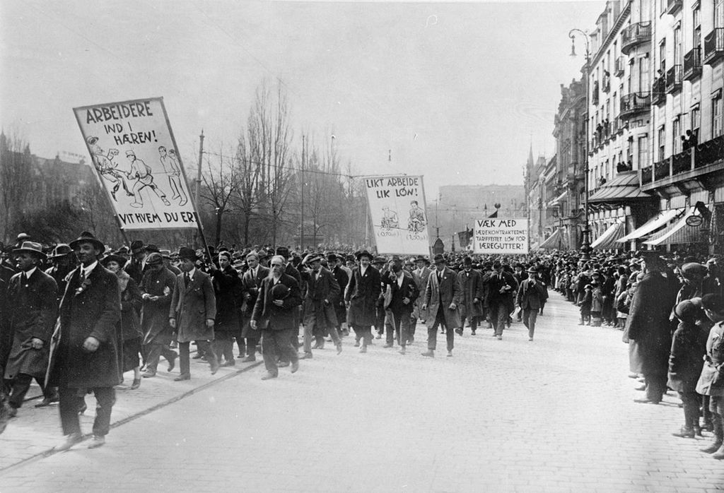 Første mai-toget på Karl Johan. En av parolene har teksten: "Likt arbeide lik løn" og viser en tegning av en kvinne og en mann som gjør samme arbeid, mens mannen får over dobbelt så godt betalt.  Bare menn går i toget. Det er mange tilskuere. Foto. 