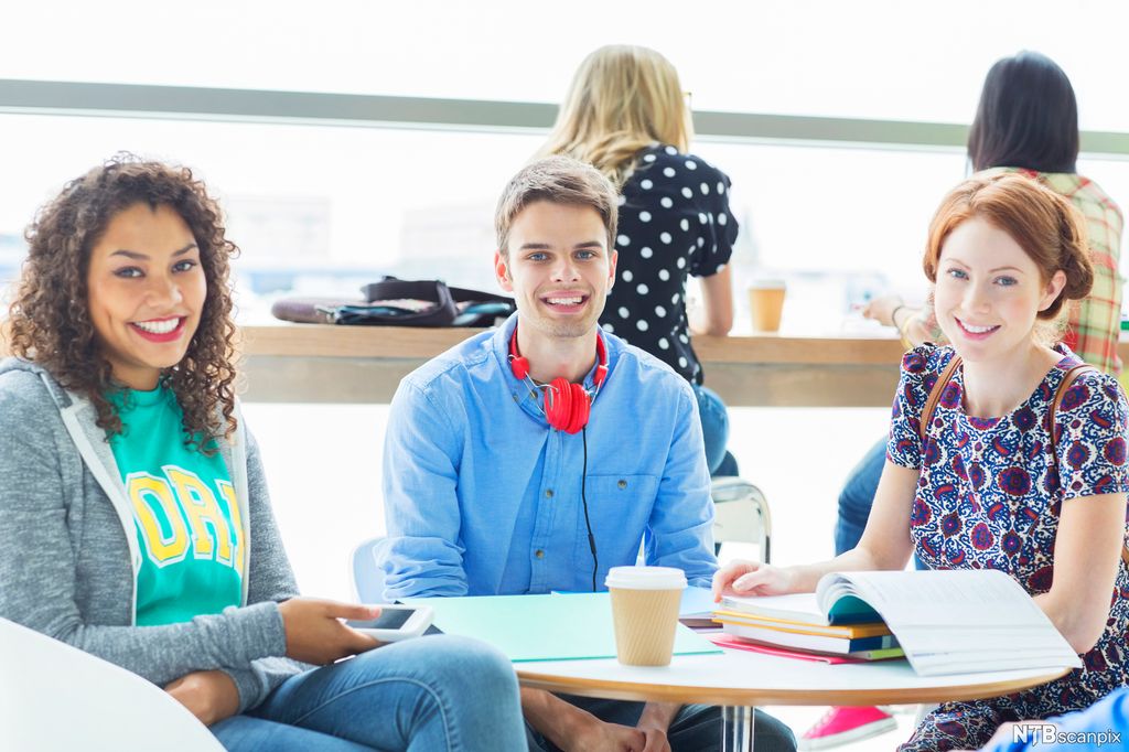 Tre ungdommer rundt et kantinebord med bøker og en pappkrus. Foto.