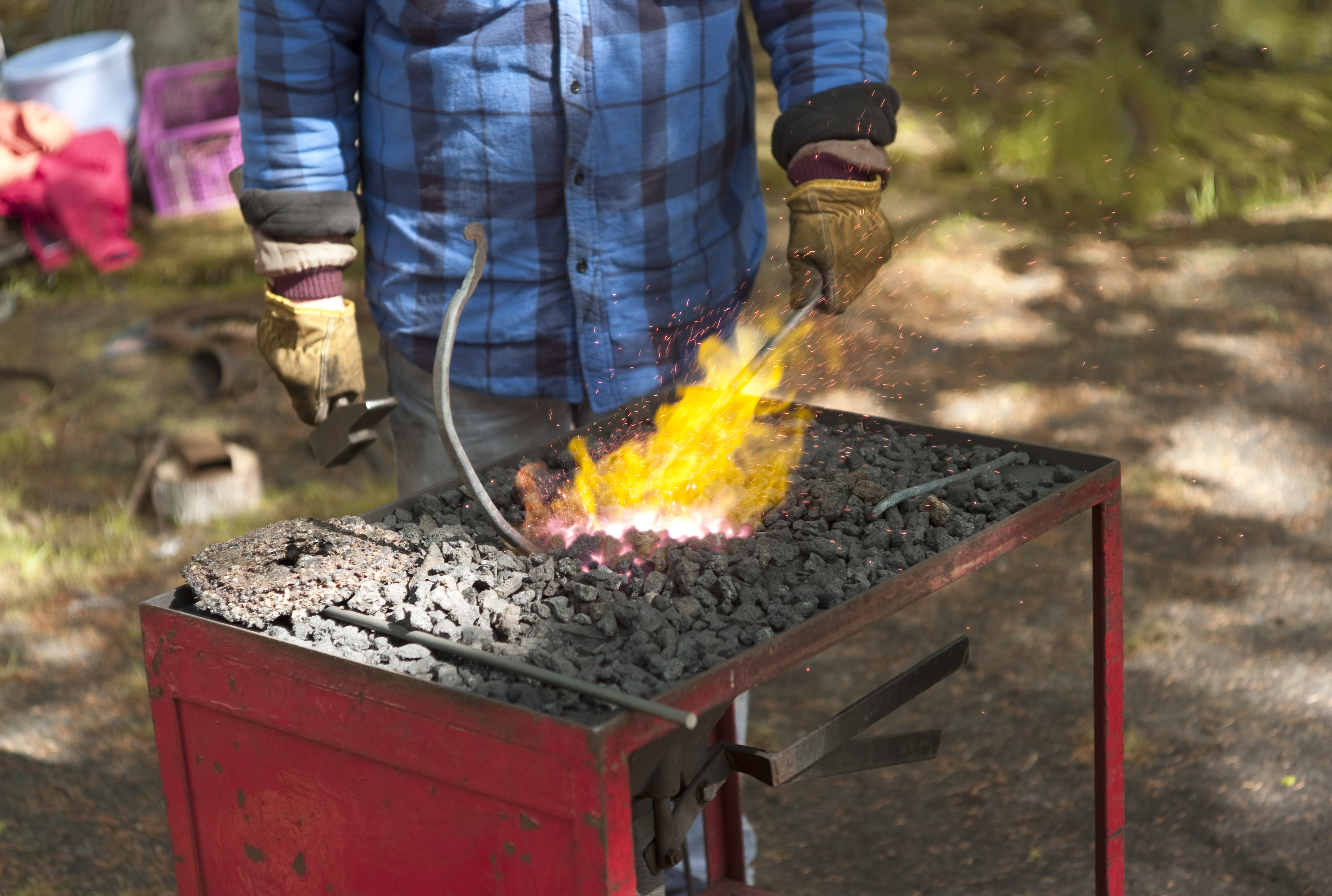 Et firkanta metallbord der bordflata er fylt med svart kull. Midt på bordet er kullet glødende, og det står opp flammer fra kullet. En person som har på seg vernehansker, holder en stålbit ned i det rødglødende kullet. Foto.