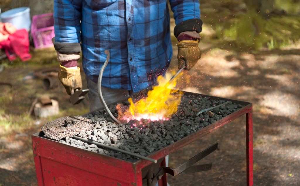 Eit firkanta metallbord der bordflata er fylt med svart kol. Midt på bordet er kolet glødande, og det står opp flammar frå kolet. Ein person som har på seg vernehanskar, held ein stålbit ned i det raudglødande kolet. Foto.
