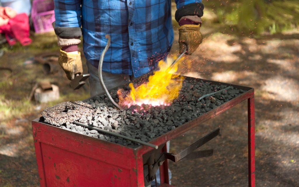 Et metallbord der overflata er fylt med kull. Kullet midt på bordet er rødglødende og varmt. En person holder en stålbit ned i det glødende kullet. Foto.