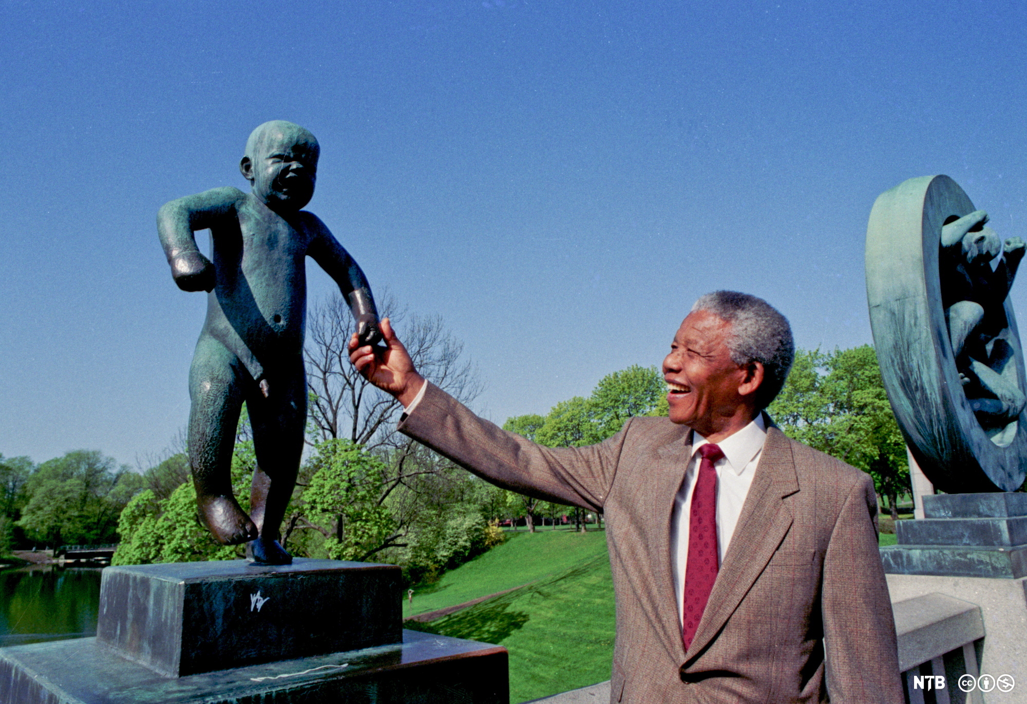 Photo: Nelson Mandela holds the hand of Gustav Vigeland's sculpture 'Sinnataggen' in Vigelandsparken in Oslo. He is smiling. He is wearing a brown suit and a red tie. 