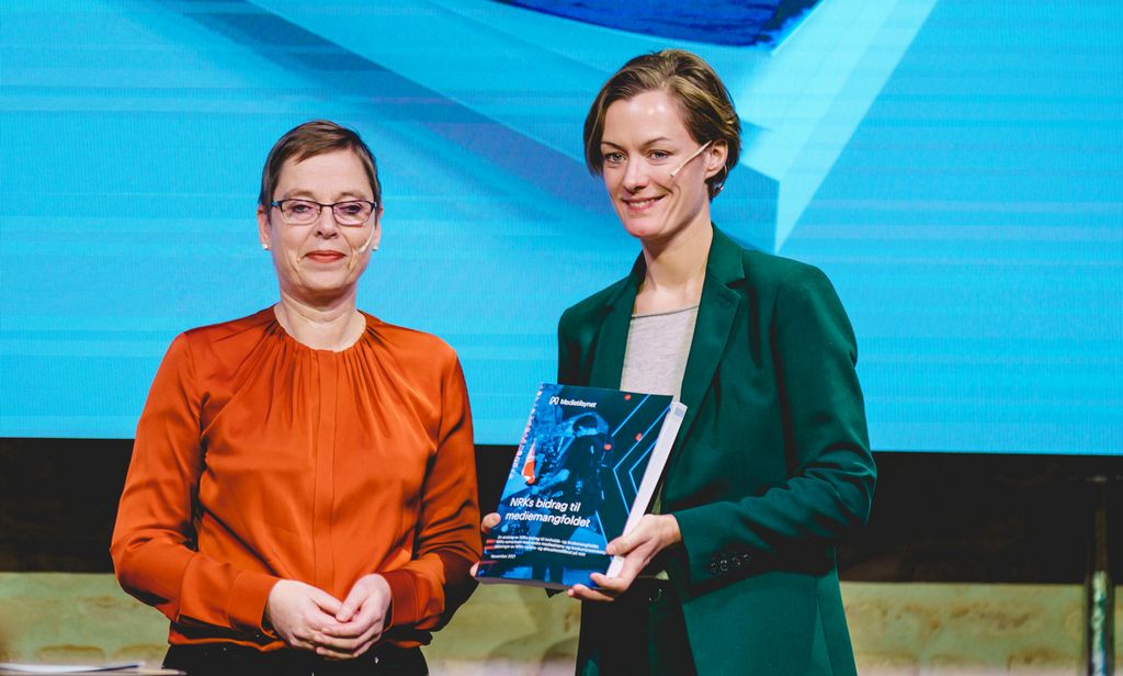 En middelaldrende kvinne i oransje bluse står sammen med en litt yngre kvinne i grønn blazer som viser fram en rapport. Foto.