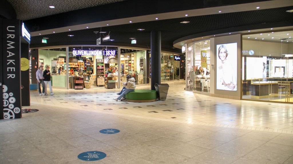 Fellesarealene på Lagunen Storsenter i Bergen. Vi ser inngangspartiet til en urmaker, en optikk butikk og en olivenbutikk. Fotografi.