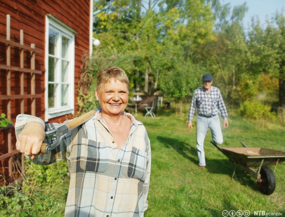Eldre kvinne står ute i hagen. Hun holder en spade over skuldra og smiler. Foto.