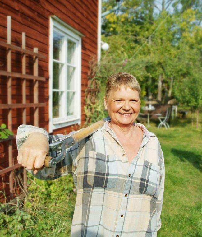 Eldre kvinne står ute i hagen. Hun holder en spade over skuldra og smiler. Foto.