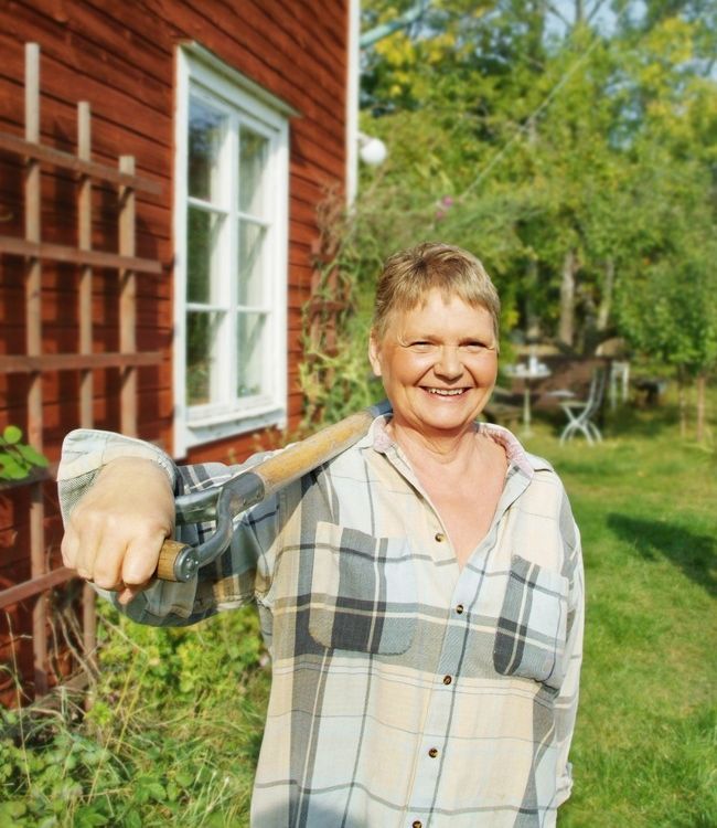 Eldre kvinne står ute i hagen. Ho held ein spade over skuldra og smiler. Foto.