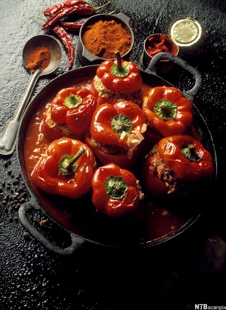 En stekepanne med sju paprikaer. Hver paprika er delt og fylt med en kjøttaktig masse. Ved siden av stekepanna er det to røde chili og tre små skåler med ulike typer krydder. Foto.