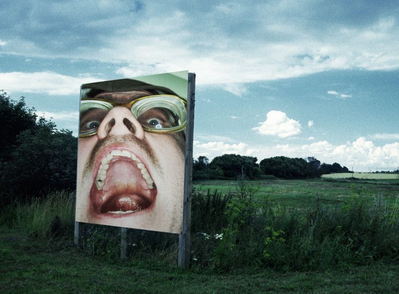 Reklameplakat i åpent landskap. Den viser en mann som gaper opp, slik at ganen hans vises. Foto.