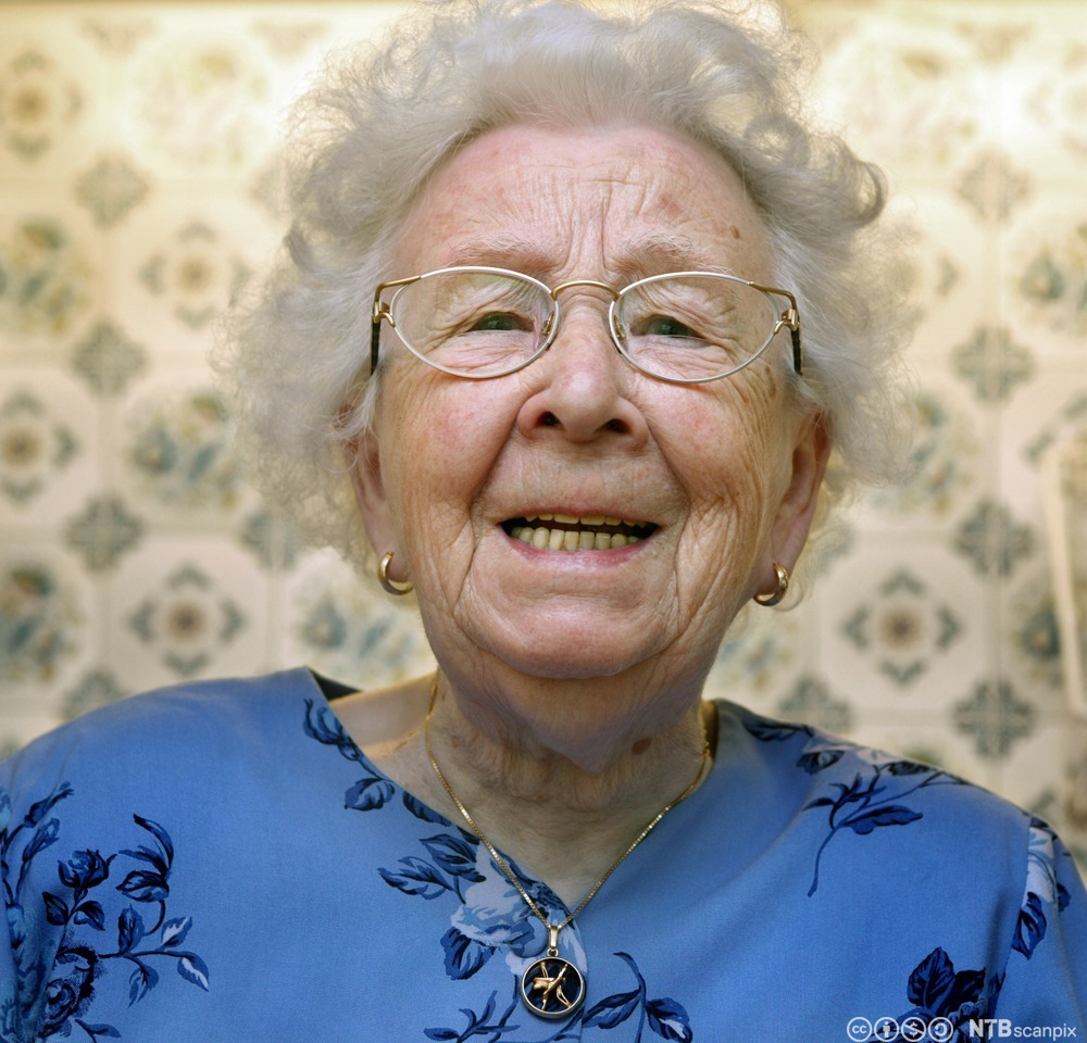 Portrett av smilende gammel kvinne med blå kjole, hvitt hår og briller. Foto.