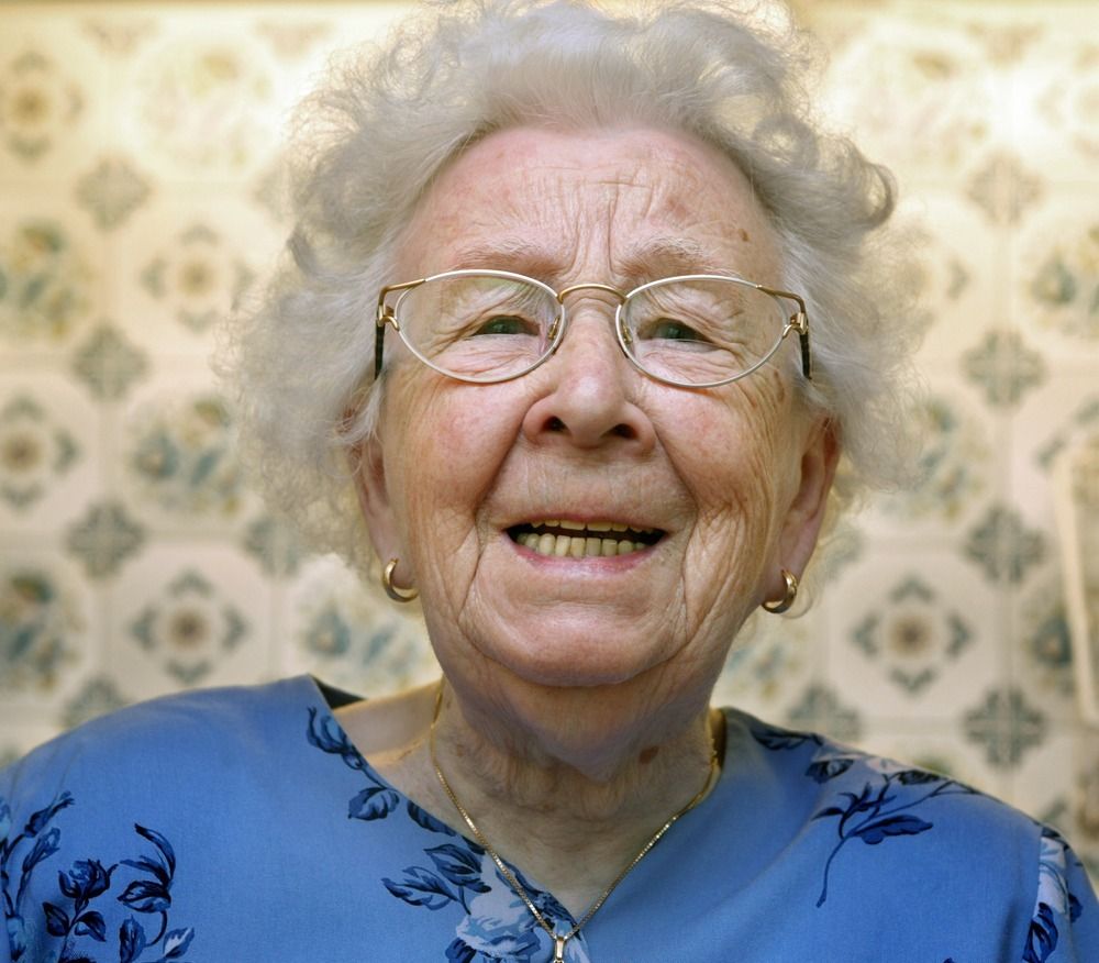 Portrett av smilende gammel kvinne med blå kjole, hvitt hår og briller. Foto.