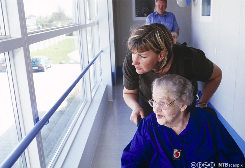 Eldre dame i rullestol og en pleier ser ut av vinduet. Foto.