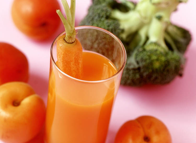 Foto av et glass med gulrotjus. I bakgrunnen ligger brokkoli og frukt.