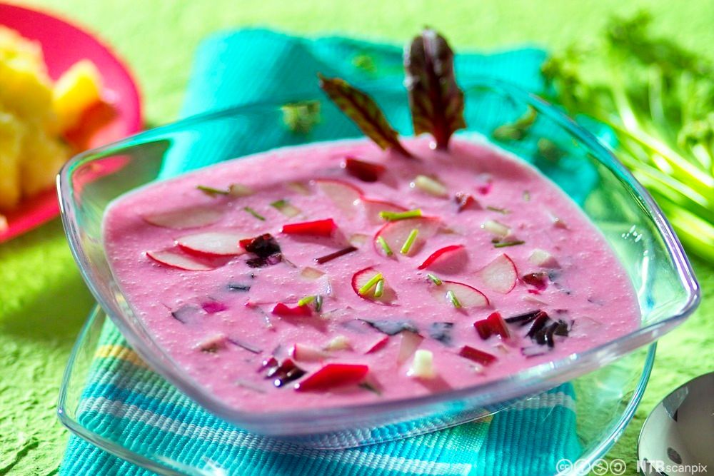 Rosa rødbetsuppe i en glasstallerken. Flere råvarer i mange farger stikker opp gjennom overflaten. Foto.