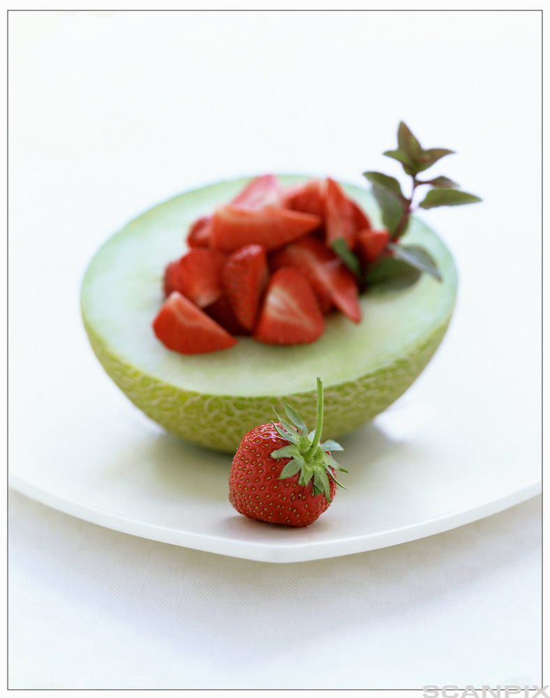 Halv grønn melon anrettet på et hvitt fat. Frøene i kjernen er fjernet og erstattet med oppskårne jordbær. Foto.