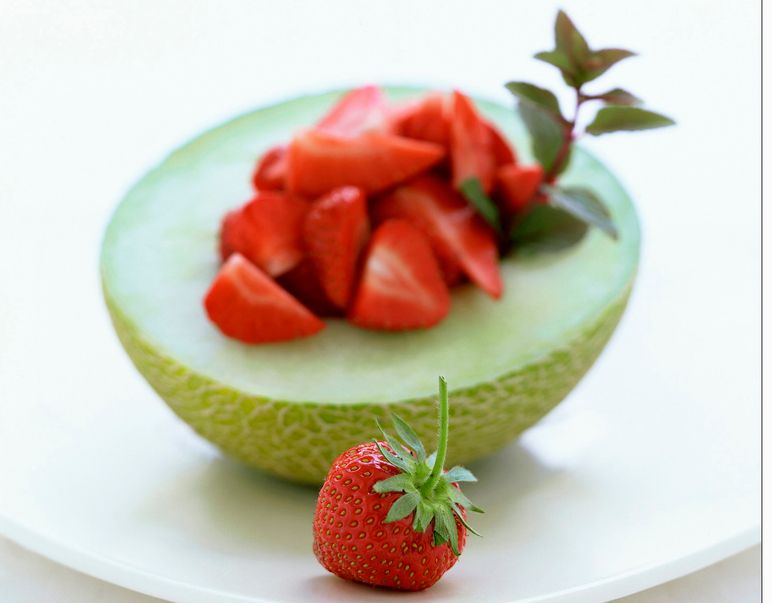 Halv grønn melon anrettet på et hvitt fat. Frøene i kjernen er fjernet og erstattet med oppskårne jordbær. Foto.