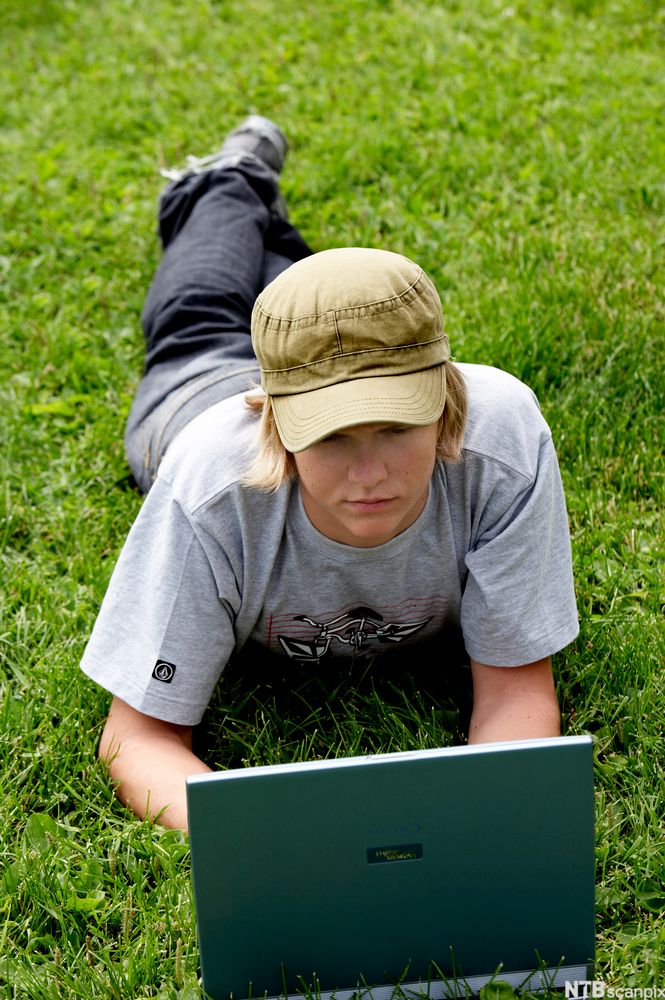 Gutt ligger i gresset med laptop. Foto.