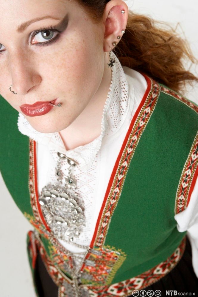 Jente i bunad med grønn vest og søljer, som er tungt sminket og har piercing i leppe, nese og øre. Foto.